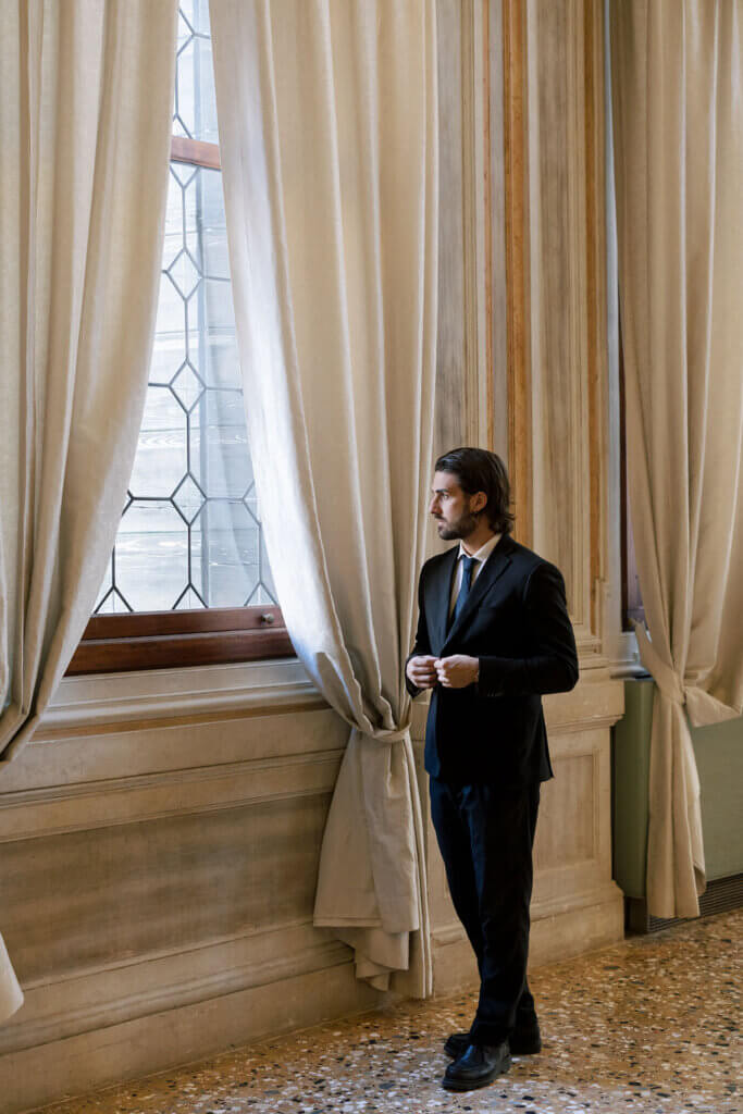 The groom looks out the window of the ballroom at an Italy Destination Wedding in Venice.