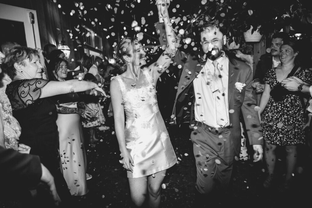 A black and white shot of the newly married couple, arms raised high as they exit their reception while confetti falls around them at Riverdale Manor in Lancaster, Pennsylvania.