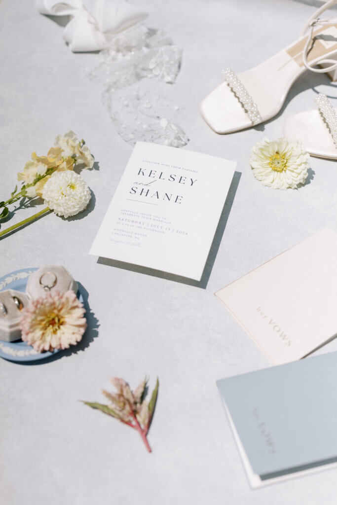 A flat lay detailing the bride and groom's details, including their rings, vow books, her heels, and their invitation at Riverdale Manor in Lancaster, Pennsylvania.