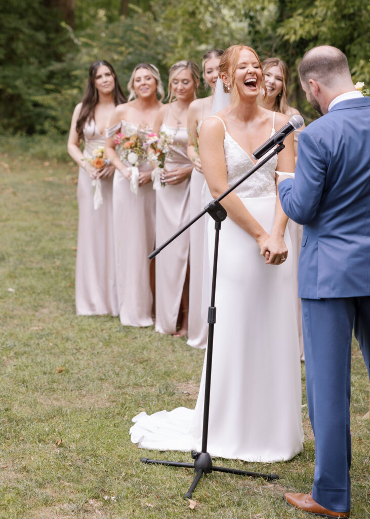 The bride laughs as the groom reads his vows with the bridesmaids looking on in the background at their outdoor ceremony at Riverdale Manor in Lancaster, Pennsylvania.