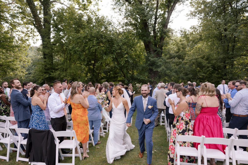 The bride and groom walk hand-in-hand down the aisle after their outdoor ceremony while the guests stand and clap at Riverdale Manor in Lancaster, Pennsylvania.