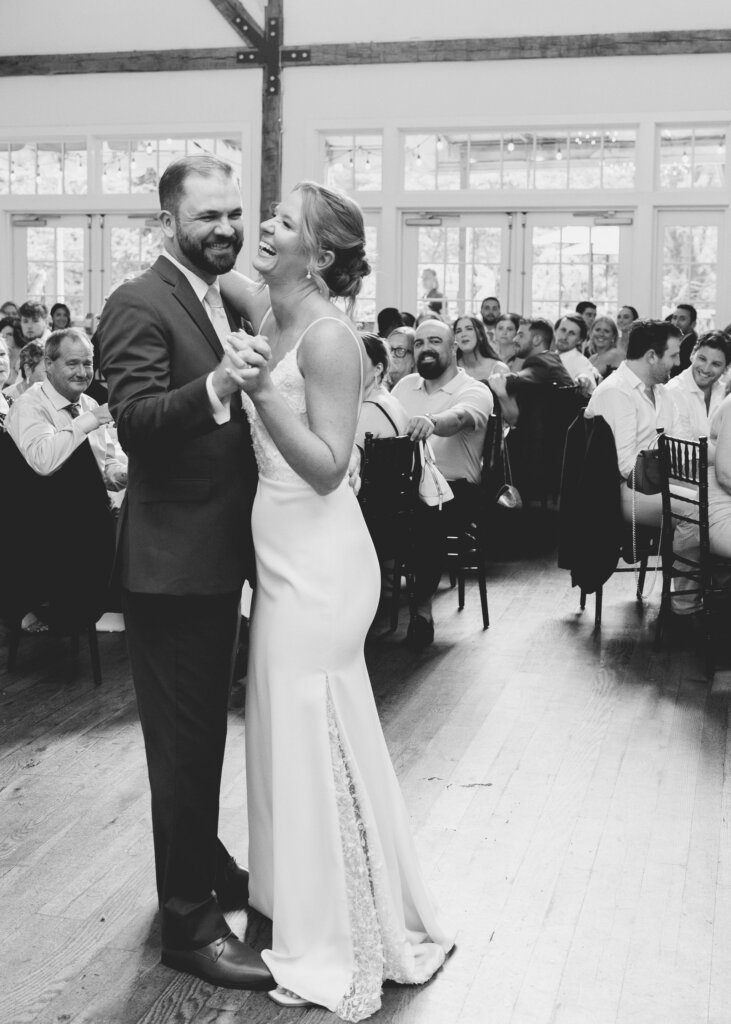 A black and white candid shot of the bride and groom laughing as they enjoy their first dance as a married couple while the guests look on at their Riverdale Manor wedding in Lancaster, Pennsylvania.