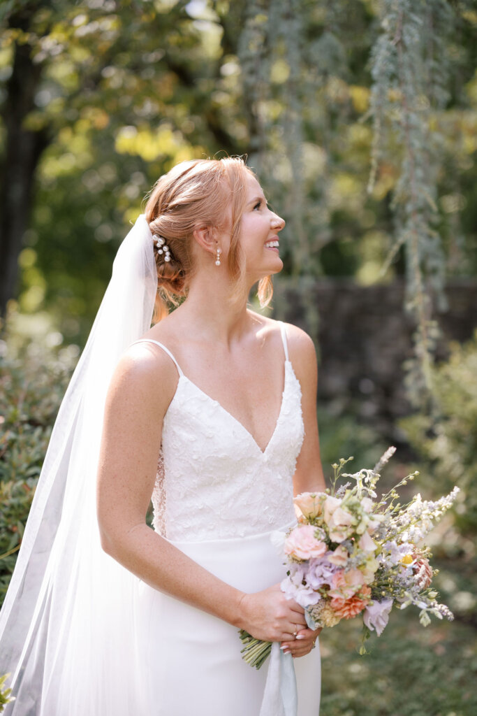 A portrait of a bride holding her bouquet, smiling over her shoulder in the afternoon sunlight on her wedding day at Riverdale Manor in Lancaster, Pennsylvania.