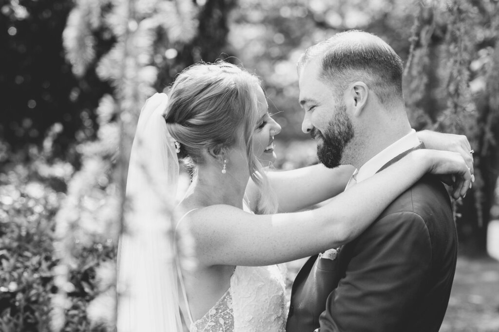 A bride and groom laugh together in a black and white portrait with her arms draped over his shoulders at Riverdale Manor in Lancaster, Pennsylvania.