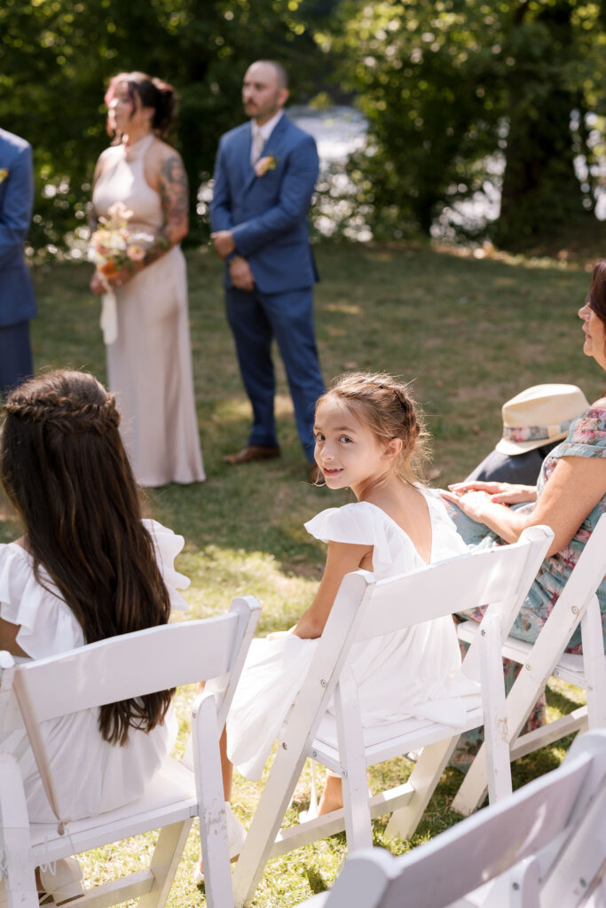 A flower girl smiles while seated front-row at an outdoor wedding ceremony at Riverdale Manor in Lancaster, Pennsylvania.