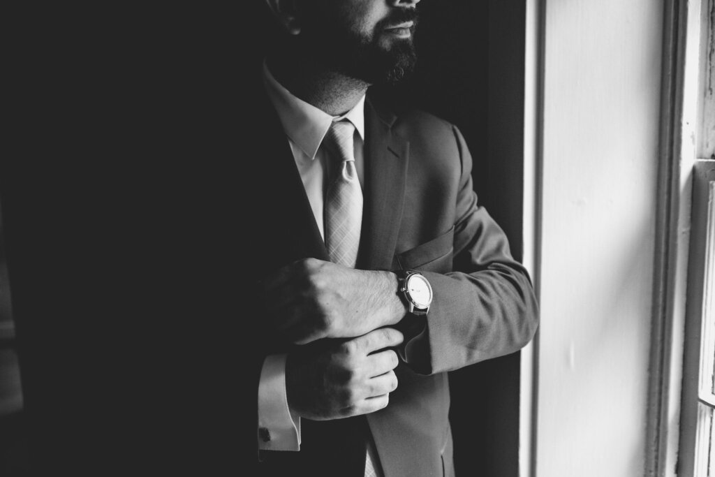 A black and white image of the groom adjusting his cuffs while gazing out the window before his wedding at Riverdale Manor in Lancaster, Pennsylvania.